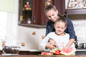 Cute little girl and her beautiful mom are cutting fruits, red watermelon and smiling while cooking in kitchen at home. Happy Family