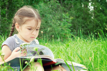 A girl in summer sits on the grass and reads a book outdoors