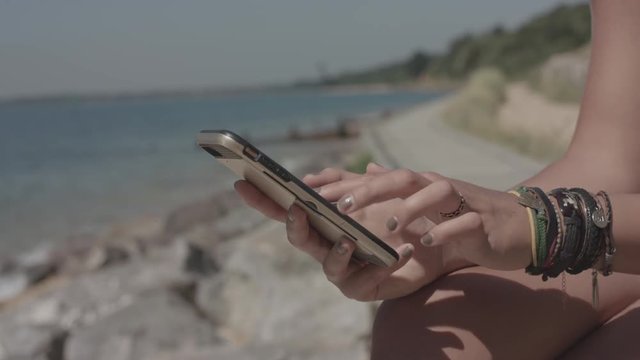 Young Attractive Woman Sat On The Beach Uses Her Smart Phone To Make Online Purchase In Slowmotion - Ungraded