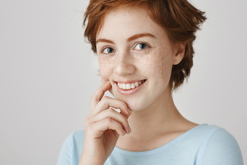 I am curious with your offer. Studio portrait of cute charismatic european ginger girl with freckles smiling broadly while scratching cheek with index finger, hesitating to give reply at once