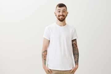 Charming tattooed carefree boyfriend with fair hair and moustache holding hands in pockets and smiling joyfully, feeling happy receiving paycheck, wanting celebrate, standing casually over gray wall