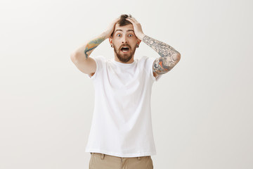 Guy heard horrible news, being very shocked. Portrait of nervous stunned emotive bearded man in white t-shirt, holding hands on head and screaming from fear, standing over gray background