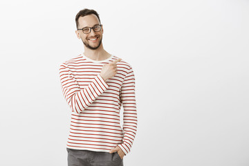 Portrat of proud confident aduly guy in glasses, holding hand in pocket and pointing at upper right corner with broad smile on face, being satisfied and joyful, feeling happy over gray background