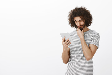 Portrait of pleased curious attractive guy with beard and afro haircut, rubbing chin while gazing at digital tablet, thinking or considering, weighing options over gray background