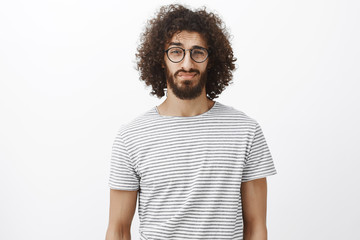 Fototapeta na wymiar Studio shot of unsure good-looking bearded male with curly hair in trendy black glasses, lifting eyebrown and looking with doubt at camera, being displeased over gray background
