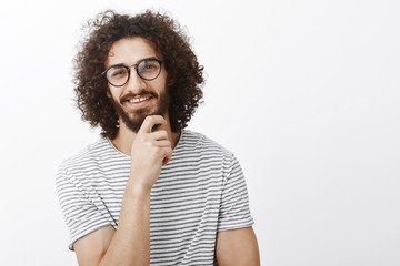 Fototapeta na wymiar Nice idea, you intrigued me. Portrait of interested smart handsome adult man with beard and curly hair in trendy glasses, smiling curiously and rubbing chin while having great thought over gray wall