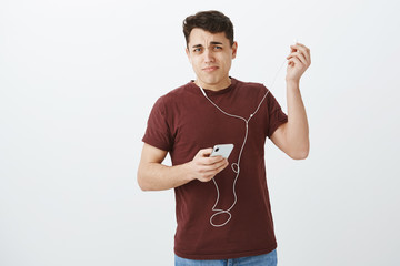 Portrait of displeased shocked attractive man in casual outfit, putting out earphones with disliking grimace, holding smartphone, being surprised with loud noise in earbud over gray wall