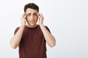 Portrait of unhappy european guy in red t-shirt, touchign temples with fingers and frowning, suffering from migraine or headache, standing displeased and gloomy over gray background