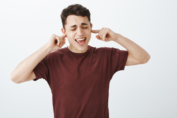 Fototapeta na wymiar Portrait of good-looking childish caucasian guy in red t-shirt, yelling with closed eyes and covering ears with index fingers, not wanting hear girlfriend during fight, standing over gray background