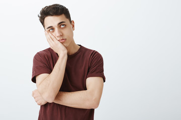 Portrait of impatient bored attractive guy in ordinary red t-shirt, leaning face on palm and looking up, waiting for end of time, being tired and feeling boredom while standing over gray background