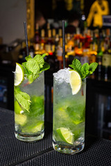 Two Mojito cocktails on a bar counter on blurred background with bottles.