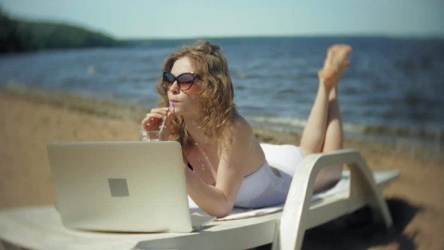 A young girl in a white bikini lies and tans on a deckchair on a sea sandy beach and is working on a laptop