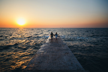 Young couple running at the pier towards the sunrise sun at the beach summer time