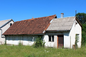 Very small abandoned houses with dilapidated facade and roof tiles, windows and doors covered with old wooden blinds surrounded with high uncut grass, flowers, trees and other vegetation on clear blue