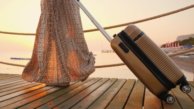 A woman with a bag on wheels rides the pier towards the sea, the rising sun beautifully illuminates her dress. In the frame, only the legs are visible. Go on a cruise concept