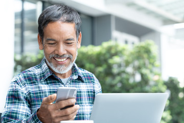 Smiling happy mature man with white stylish short beard using smartphone gadget serving internet at coffee shop cafe outdoor. Laughing old man using social media network technology feeling excited.
