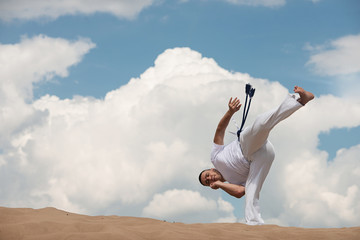 Young guy trains capoeira on sky backround. A men performs martial the kick