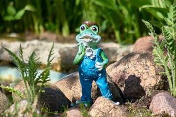 Decorative frog in the garden on the background of stones. Toy frog in overalls wipes his face with...
