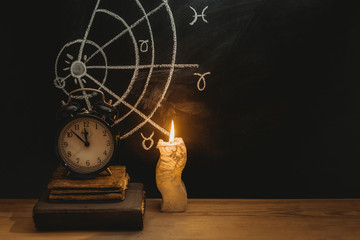 Alarm clock and old books next to a candle on a horoscope background drawn on a chalkboard