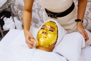 Beauty clinic staff helping young woman getting 24 karat gold facial treatment at the beauty clinic. The treatment of using real gold for youthful skin.