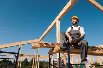 The man builder sits on the edge of the roof of the frame house, in a yellow helmet and gray overalls. The blue sky and clear sunny day.