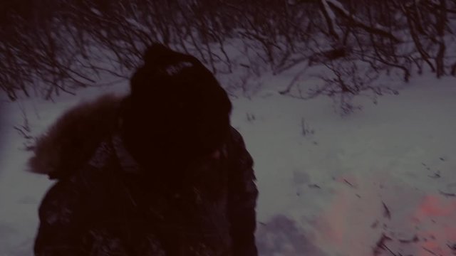 Cree man warming food and tea in winter over fire in snow, adding wood, northern Quebec