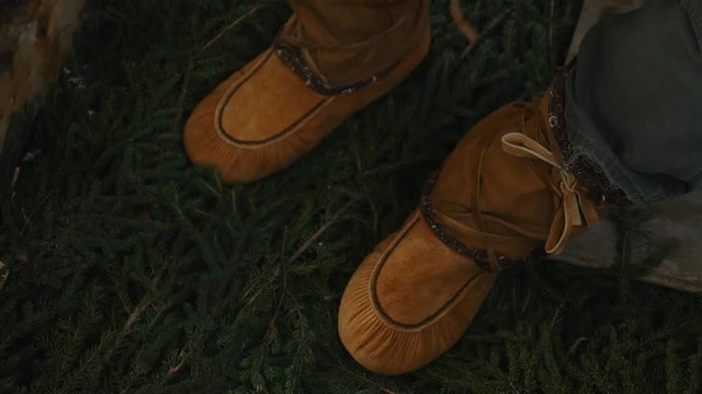 Man tying traditional Cree winter mukluks on spruce boughs, inside tent