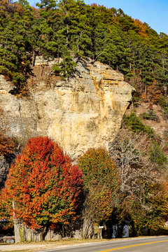 Sheer rock bluff in the Autumn with pinen trees growing on top and trees with colored foliage and a two lane highway and mailbox below