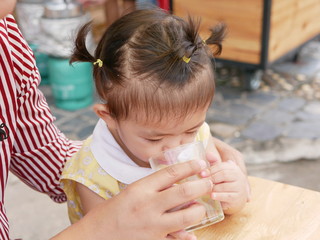 Obraz premium Little Asian baby girl, 16 months old, learning to drink directly from a glass / cup with help from her mother