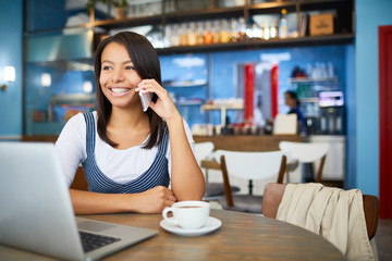 Smiling businesswoman in casualwear sitting by table in cafe, having drink and phoning to someone