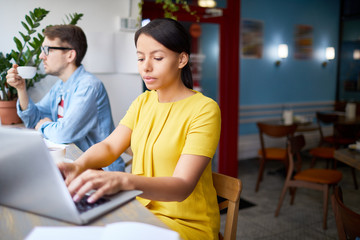 Obraz premium Serious businesswoman typing on laptop keypad while sitting in cafe with colleague near by