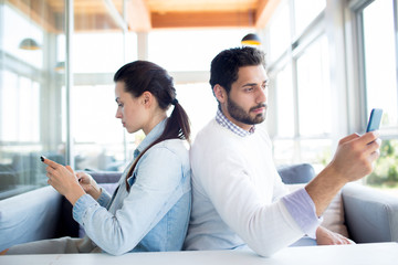 Young casual man and woman sitting back to back in cafe and communicating in their smartphones