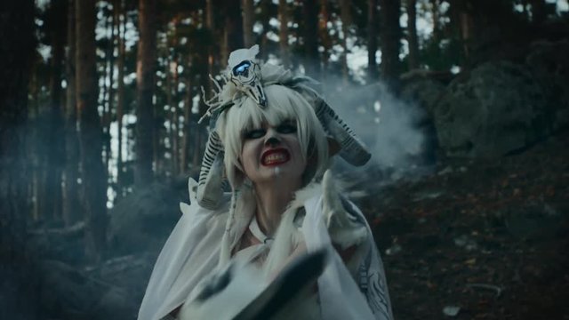 Terrifying female faun is waving by huge axe in forest. She is looking at camera in darkness, aggressive and scary