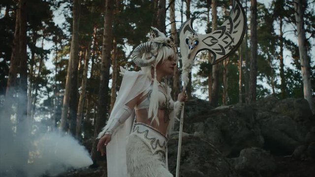 Actrees walking across the forest in a white costume of a mystical creature carrying a giant axe. Costume with white fur, magical evening in forest.