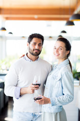 Cheerful and amorous couple with glasses of red wine looking at you while celebrating anniversary in restaurant
