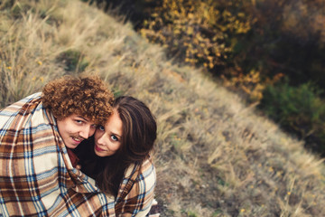 Loving cute couple sitting on grass and hiding behind rug in autumn.