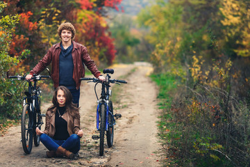 Obraz premium curly-haired mustachioed guy and an ore-haired girl in autumn stand with bicycles on road and show cheerful emotions.