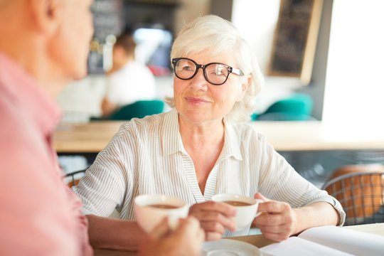 Aged woman with cup of tea looking at her colleague during discussion of ideas at start-up meeting