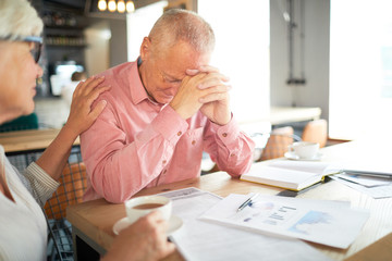 Aged frustrated businessman touching his forehead while leaning over papers on table and his...