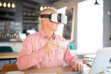 Senior man in vr goggles having tea or coffe in cafe while sitting by table in front of laptop
