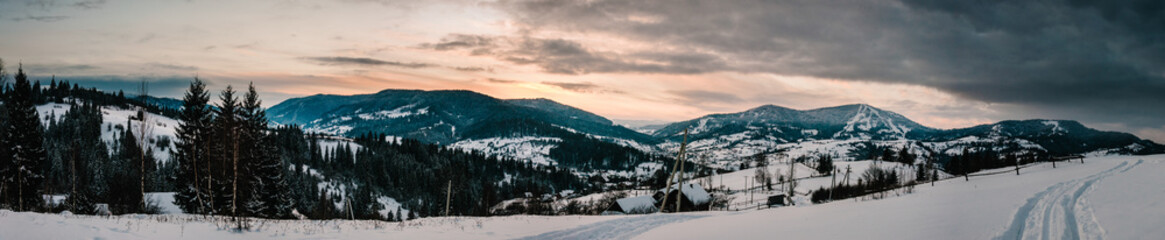 Snow-capped houses in mountains Carpathians Ukraine. On background Christmas tree in forest. Winter nature. Landscape. Top side view. Long edge. Panorama. Bukovel. Beautiful sunset over a wide valley.