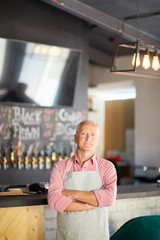 Aged waiter of cafe or restaurant with his arms crossed on chest standing by bar counter