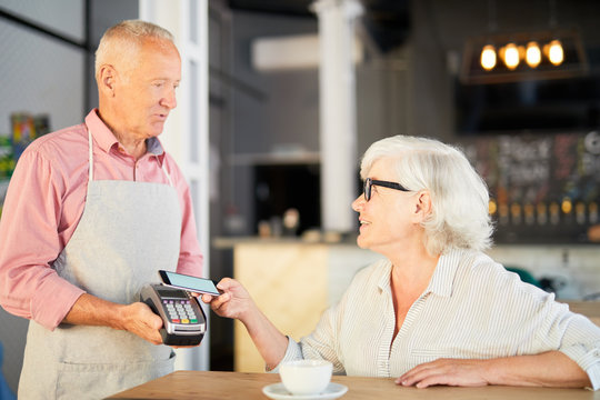 Senior waiter with payment machine talking to client paying for order through smartphone - Powered by Adobe