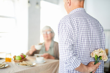 Senior man hiding bunch of beige carnations behind his back to make surprise for wife