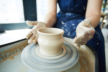 Female potter sitting by pottery wheel with her hands around rotating half-made jug