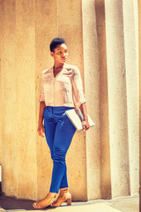 Portrait of Young African American Businesswoman in New York, with short afro hair, wearing light color V neck shirt, blue pants, brown sandal high heels, holding laptop computer, standing by column..