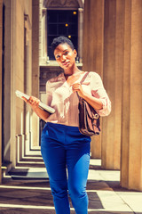 Young African American Woman in New York, with short afro hair, wearing light color V neck shirt, blue pants, shoulder carrying small bag, holding laptop computer, walking inside office building..