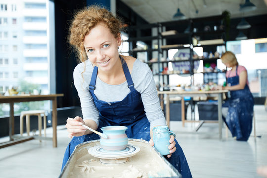 Young woman in workwear painting rotating clay jug in blue color while sitting by pottery wheel in workshop