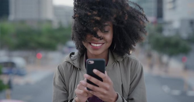 portrait of happy african american woman using smartphone texting in city smiling enjoying browsing online messages on mobile phone wind blowing afro hairstyle