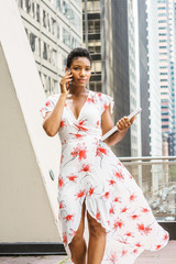 Young African American Businesswoman with short afro hair, wearing long dress, holding laptop computer, talking on cell phone, walking on balcony in New York. Street with hight buildings on background